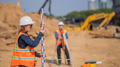Woman and man in orange safety vests and hard hats doing surveying work at road construction site.