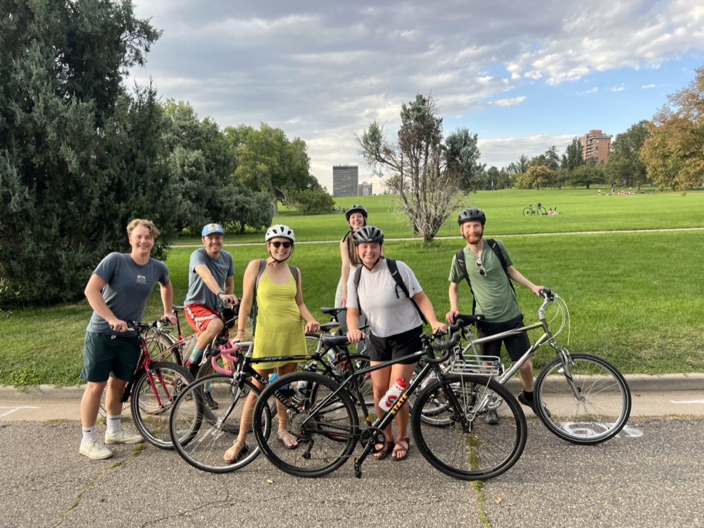 Photo of people smiling wearing helmets and holding their bikes on the side of a road in front of a feild or park