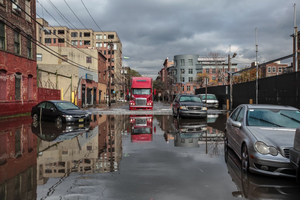 A semitruck drives down a flooded New York Street lined by cars and buildings.