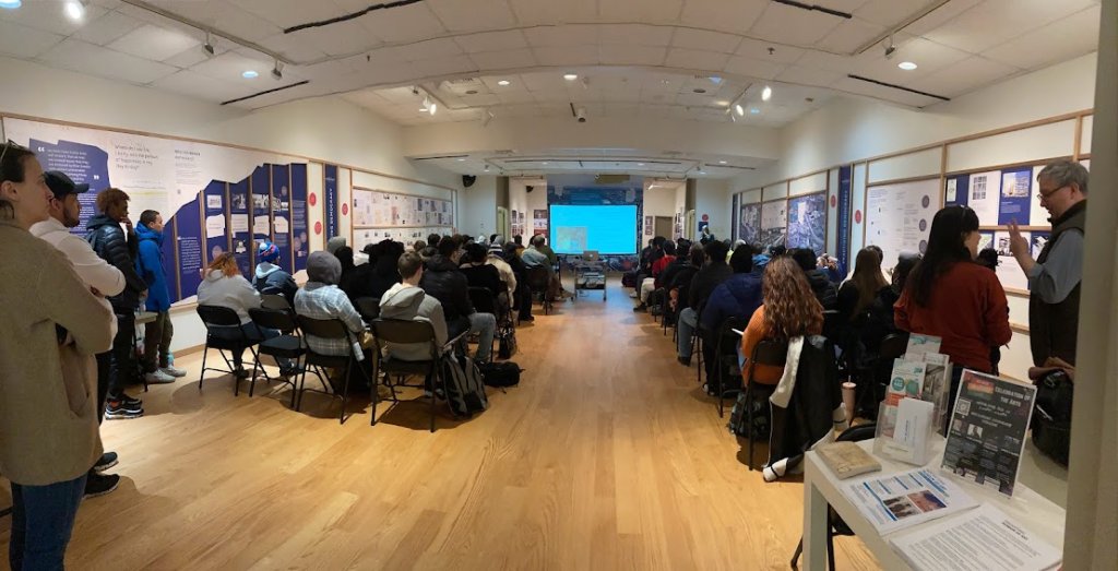 Visitors sit and stand in the foreground within the exhibition space at Housatonic Community College watching a presentation on a screen in the background.