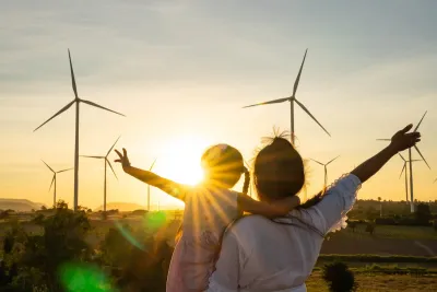 Adult holding young child facing away from camera looking at wind turbines sillhouetted against the sunset.