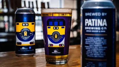Close-up of full beer glass with purple train-themed design sitting on bar between two frosty tall cans.