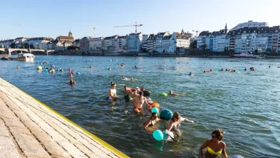 People swimming in Rhine River on a sunny day with low-rise, dense buildings along waterfront in background.