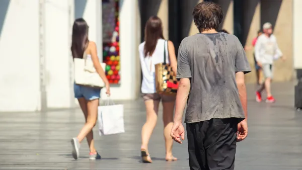 Man walking down city sidewalk with sweat on back of his t-shirt on hot day.